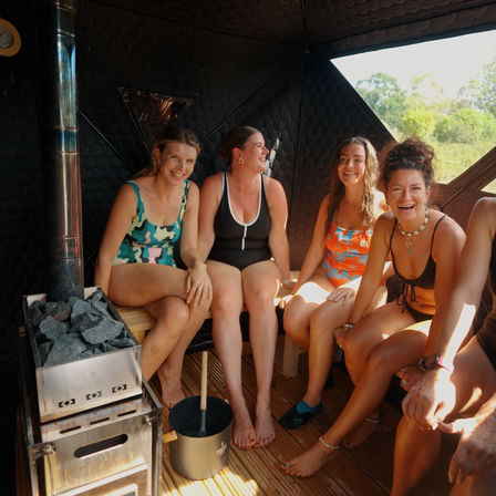 Four women in swimsuits laughing together in a sunlit wooden outdoor sauna beside a hot-stone stove with a green forest view