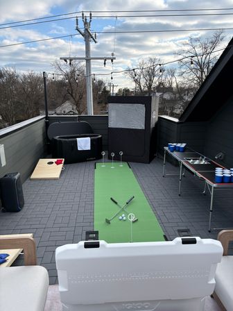 Rooftop patio deck with a green putting mat, two putters and golf balls, inflatable hot tub, portable sauna tent, cornhole board, beer-pong table with cups, string lights and overcast suburban skyline