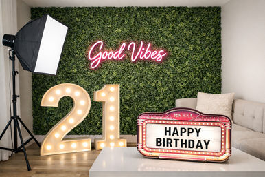 Indoor 21st birthday photo backdrop with a green foliage wall, pink neon "Good Vibes" sign, illuminated marquee numbers "21", retro "Happy Birthday" marquee, studio softbox light and beige sofa.