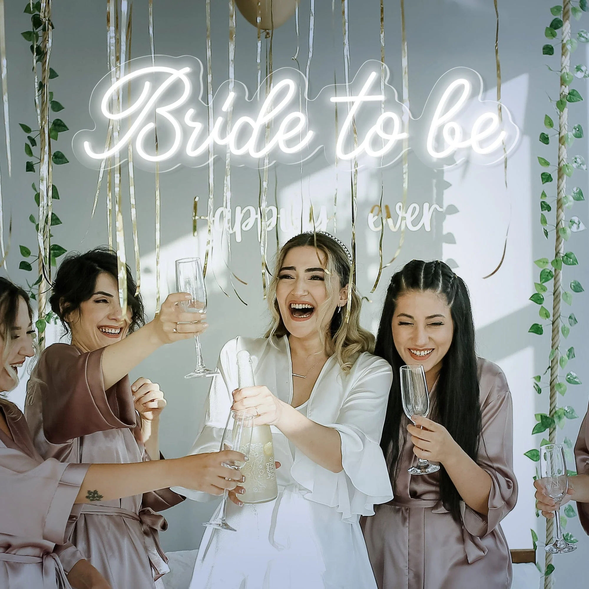 Bride-to-be and bridesmaids in satin robes laughing and toasting with champagne under a glowing "Bride to be" sign and gold streamers at a bridal shower.