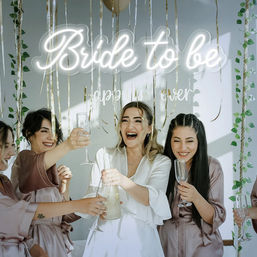 Bride-to-be and bridesmaids in satin robes laughing and toasting with champagne under a glowing "Bride to be" sign and gold streamers at a bridal shower.