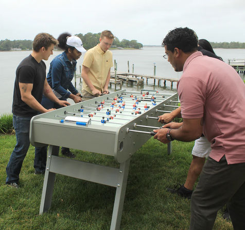 Group playing foosball at a gray outdoor table on a grassy waterfront lawn with wooden docks and calm river in the background.