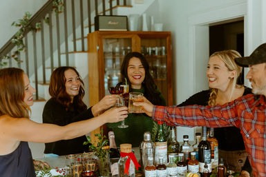 Five friends toasting cocktails at a home bar by a staircase, smiling and celebrating a cozy holiday gathering with bottles and mixers on the counter.