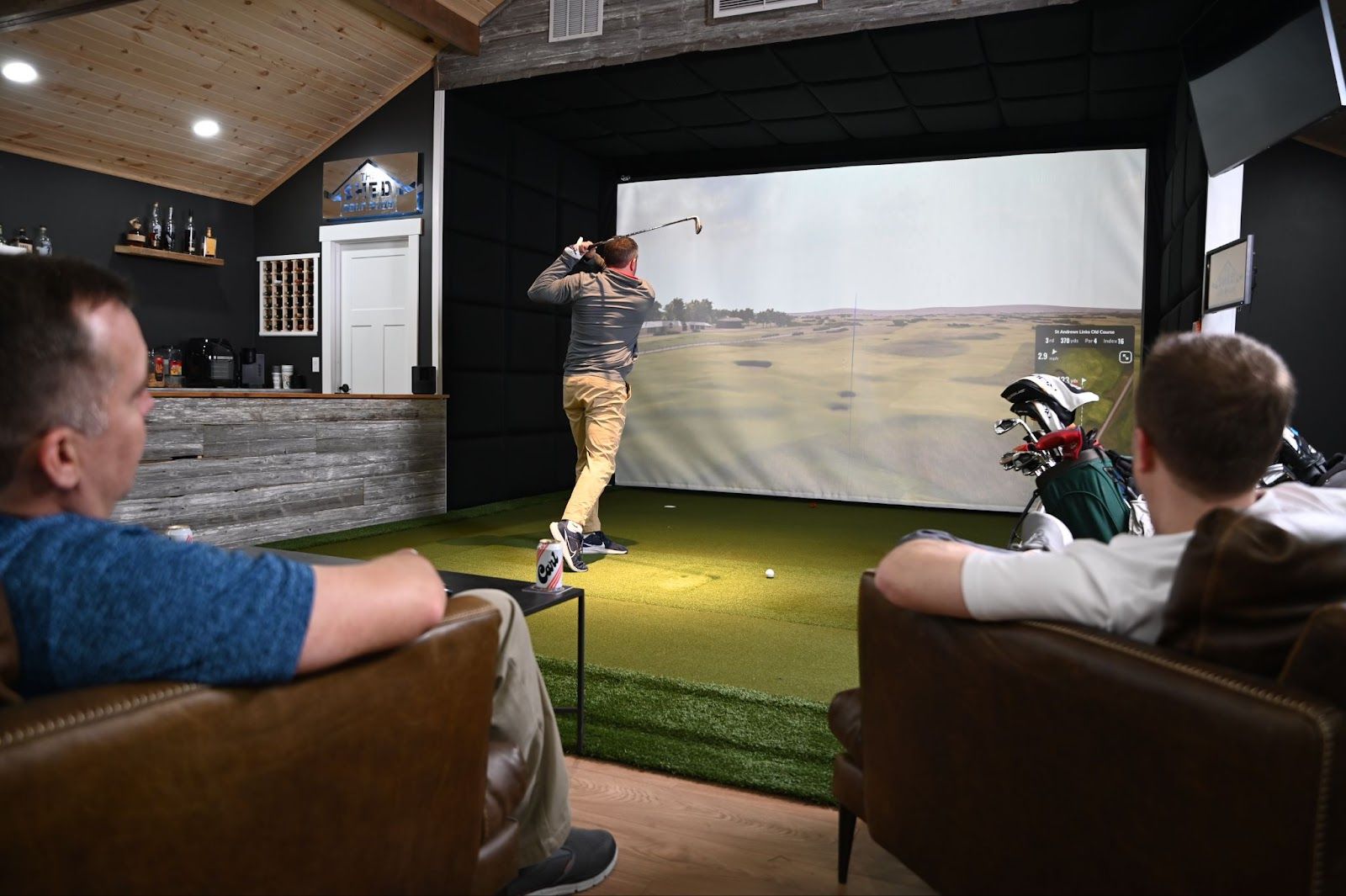 Cozy indoor golf simulator scene: golfer mid-swing toward a large projected virtual course while two friends watch from leather chairs, turf mat and golf bag nearby.