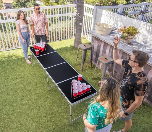 Four friends playing beer pong with red cups on a foldable black table in a sunny backyard lawn beside a white fence and outdoor bar.