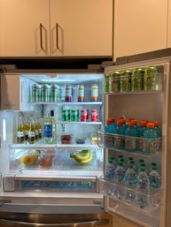 Open stainless-steel refrigerator in a modern kitchen, neatly stocked with bottled water and sports drinks, cans of soda and sparkling water, beer and wine, plus bananas and berries.