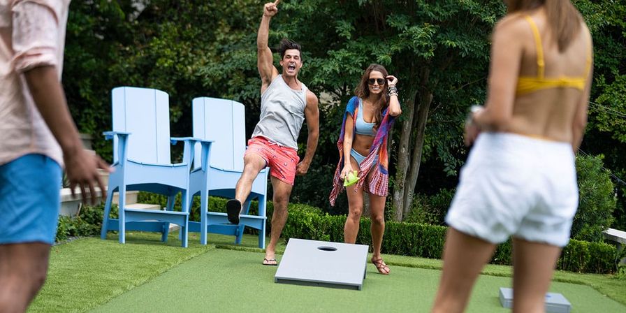 Friends playing cornhole on a sunny backyard lawn — a man cheers while a woman readies a beanbag, with blue Adirondack chairs and a leafy garden in the background.