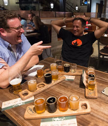 Two friends laughing at a wooden table in a cozy craft brewery taproom, sharing multiple wooden beer flight paddles with assorted pale, amber and dark beer samples, glasses, a notebook and smartphone visible.