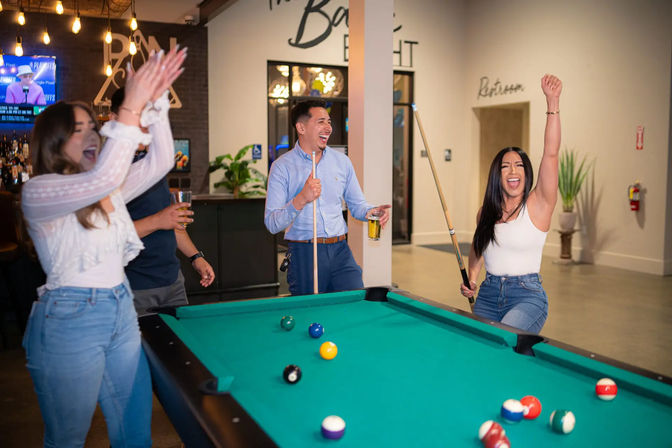 Friends celebrating a night out at a neighborhood bar pool table — woman raises cue in victory, man laughs with a beer, companion claps as billiard balls scatter on green felt.