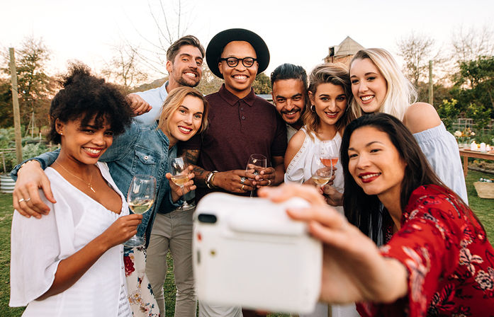 Diverse group of friends smiling and raising wine glasses while taking a selfie at an outdoor garden party at sunset.