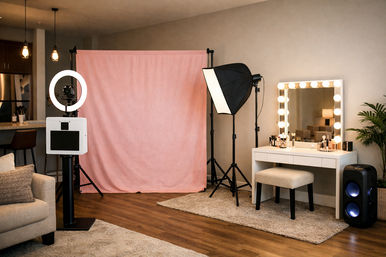 Cozy home photo studio with blush pink backdrop, ring light and camera on a stand, softbox lighting, illuminated Hollywood-style vanity with makeup and stool on hardwood floor