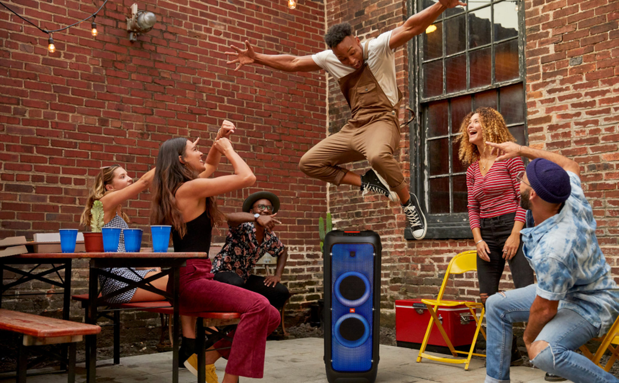 Friends partying in an urban brick-walled patio — person mid-jump as others cheer around a blue-lit portable party speaker, picnic table and string lights.