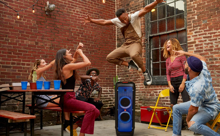 Friends partying in an urban brick-walled patio — person mid-jump as others cheer around a blue-lit portable party speaker, picnic table and string lights.