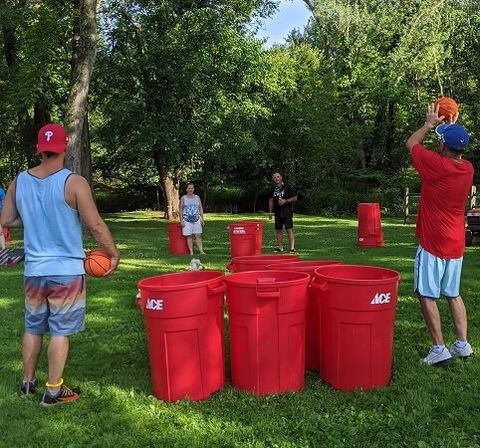 Four people playing a DIY lawn basketball game in a sunny, tree-lined park, tossing orange basketballs into grouped red plastic bins on green grass.