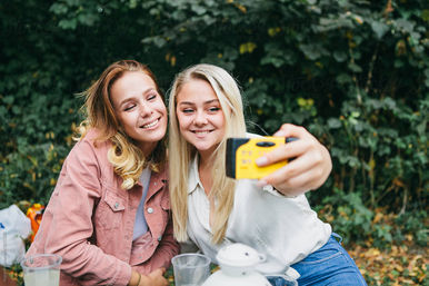 Two smiling friends take a selfie with a yellow disposable camera during an outdoor picnic in a leafy park.