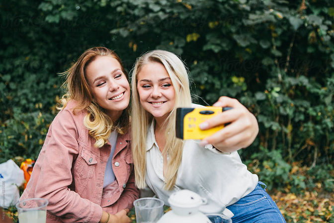 Two smiling friends take a selfie with a yellow disposable camera during an outdoor picnic in a leafy park.