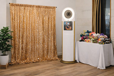 Indoor event photo booth with a shimmering gold sequin backdrop, white circular kiosk with preview screen, and a table of colorful party props on a wooden floor.