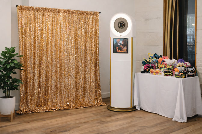 Indoor event photo booth with a shimmering gold sequin backdrop, white circular kiosk with preview screen, and a table of colorful party props on a wooden floor.