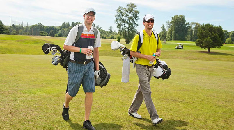 Two smiling golfers walking on a sunlit fairway carrying golf bags and drinks on a green golf course with trees and a golf cart in the background