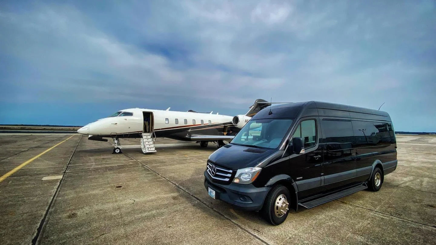 Private white business jet with open boarding stairs parked on an airport tarmac beside a black Mercedes passenger van executive shuttle under a cloudy blue sky