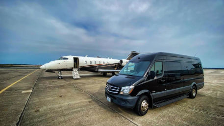 Private white business jet with open boarding stairs parked on an airport tarmac beside a black Mercedes passenger van executive shuttle under a cloudy blue sky