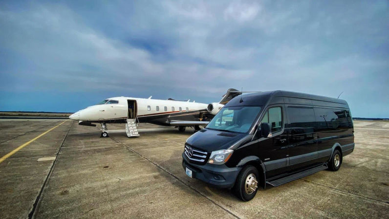 Private white business jet with open boarding stairs parked on an airport tarmac beside a black Mercedes passenger van executive shuttle under a cloudy blue sky