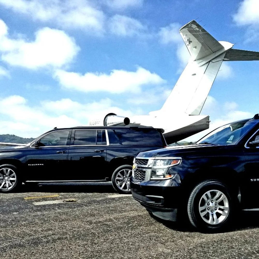Sleek black SUVs parked beside a white private jet on a sunny airport tarmac under bright blue skies — private jet ground transport scene.