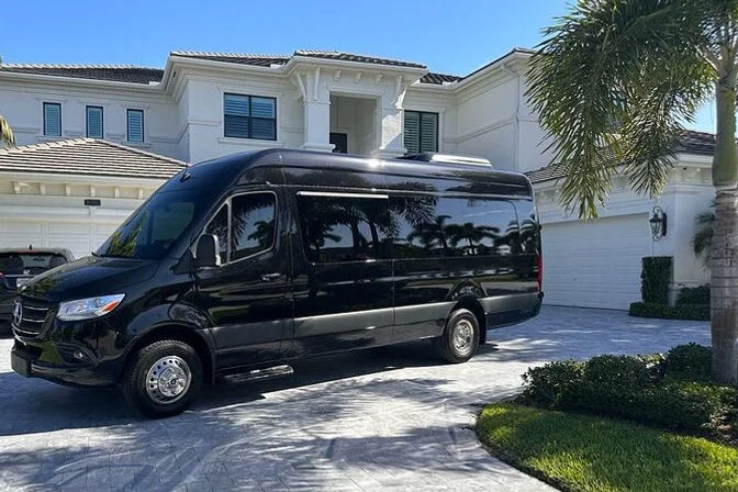 Sleek black high-roof passenger van parked in the patterned driveway of a large white two-story luxury home with palm trees under a bright blue sky.