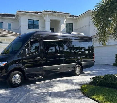 Sleek black high-roof passenger van parked in the patterned driveway of a large white two-story luxury home with palm trees under a bright blue sky.