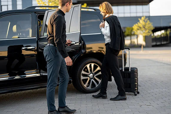 Business traveler boarding a black SUV for airport or hotel transfer while a driver opens the door; rolling suitcase on a modern urban curb