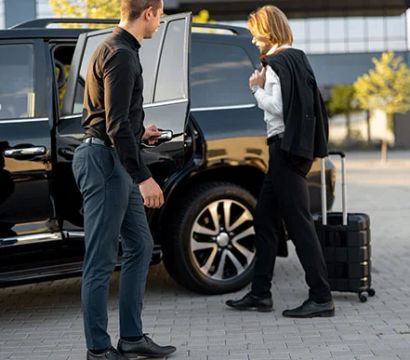 Business traveler boarding a black SUV for airport or hotel transfer while a driver opens the door; rolling suitcase on a modern urban curb