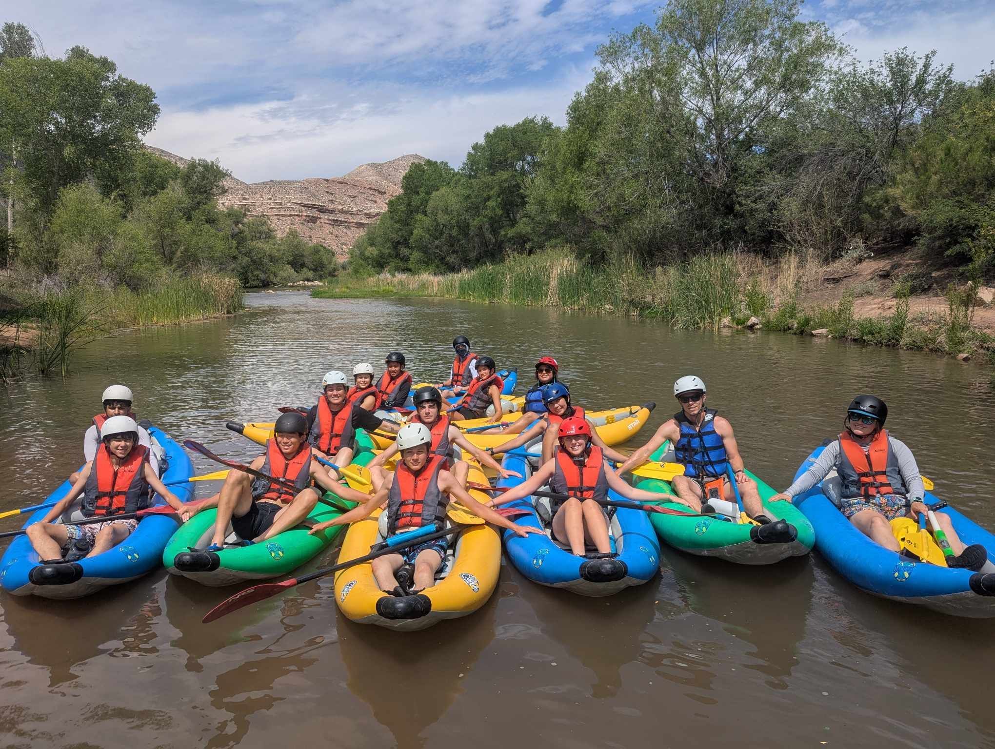 Group of smiling kayakers in colorful inflatable kayaks and life jackets paddling on a calm river surrounded by green trees and red‑rock canyon under a partly cloudy sky.