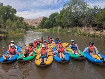 Group of smiling kayakers in colorful inflatable kayaks and life jackets paddling on a calm river surrounded by green trees and red‑rock canyon under a partly cloudy sky.