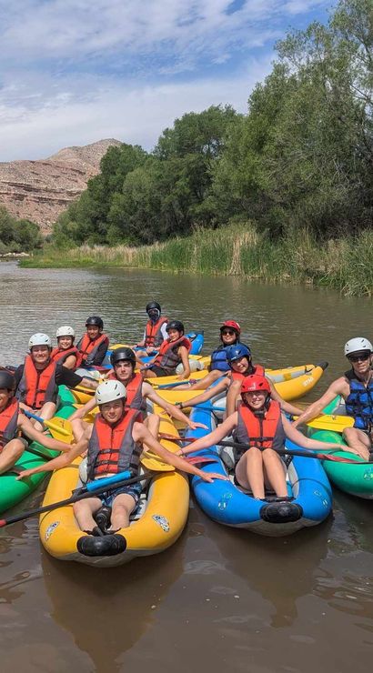 Group of smiling kayakers in colorful inflatable kayaks and life jackets paddling on a calm river surrounded by green trees and red‑rock canyon under a partly cloudy sky.