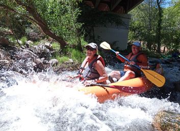 Two adults in an orange inflatable kayak wearing life jackets paddle through splashing whitewater rapids under a bridge beside a rocky, tree-lined riverbank — outdoor river rafting adventure.
