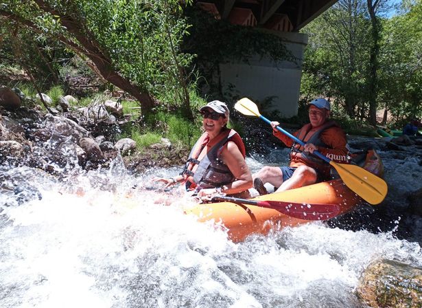 Two adults in an orange inflatable kayak wearing life jackets paddle through splashing whitewater rapids under a bridge beside a rocky, tree-lined riverbank — outdoor river rafting adventure.
