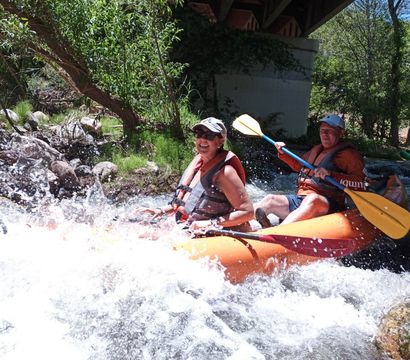 Two adults in an orange inflatable kayak wearing life jackets paddle through splashing whitewater rapids under a bridge beside a rocky, tree-lined riverbank — outdoor river rafting adventure.
