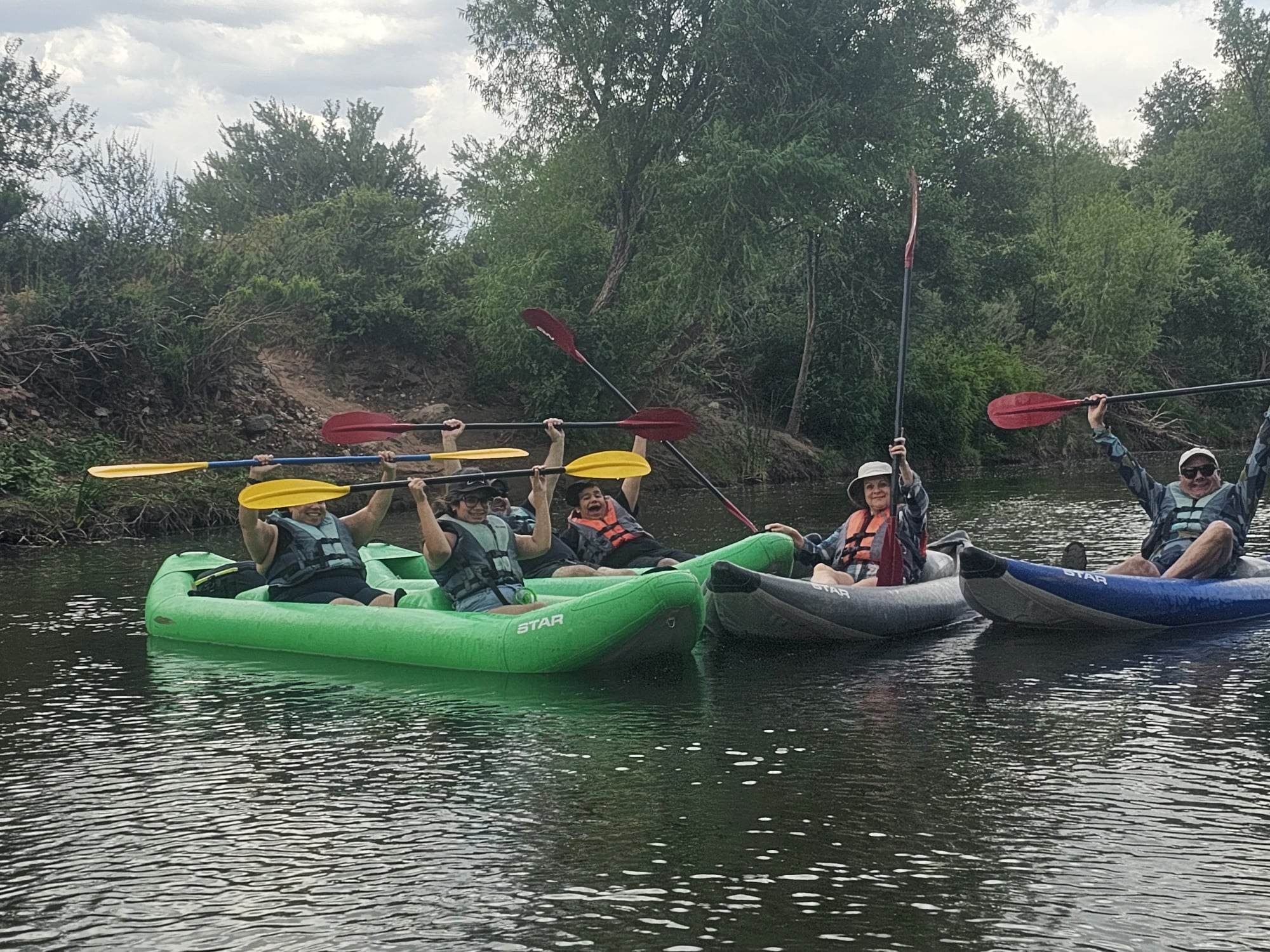 Group of kayakers in colorful inflatable kayaks holding paddles overhead on a calm river, wearing life jackets beside green trees and riverbank vegetation — outdoor kayaking adventure