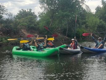 Group of kayakers in colorful inflatable kayaks holding paddles overhead on a calm river, wearing life jackets beside green trees and riverbank vegetation — outdoor kayaking adventure