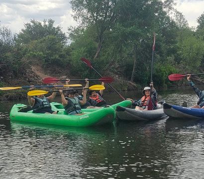 Group of kayakers in colorful inflatable kayaks holding paddles overhead on a calm river, wearing life jackets beside green trees and riverbank vegetation — outdoor kayaking adventure