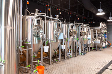 Gleaming stainless-steel conical fermentation tanks in a modern commercial brewery production room, connected by hoses, pipes and green valves on a tiled floor with an orange bucket nearby.