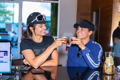 Two women clinking small beer tasting glasses at a craft brewery taproom bar counter, smiling in casual hats with a tip jar and phones nearby.