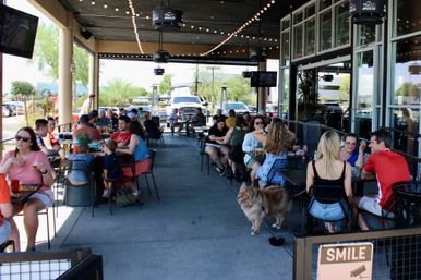 Busy dog-friendly outdoor patio dining area with string lights, groups of people eating and drinking at tables in a suburban shopping center