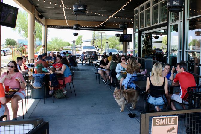 Busy dog-friendly outdoor patio dining area with string lights, groups of people eating and drinking at tables in a suburban shopping center