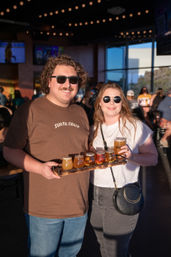 Two smiling people wearing sunglasses hold a wooden flight paddle with six colorful craft beer samples in a sunlit taproom with string lights and large windows, enjoying a relaxed brewery outing.