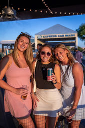 Three friends in summer dresses smiling and holding drinks under string lights at a sunlit outdoor gathering near a car wash bay.