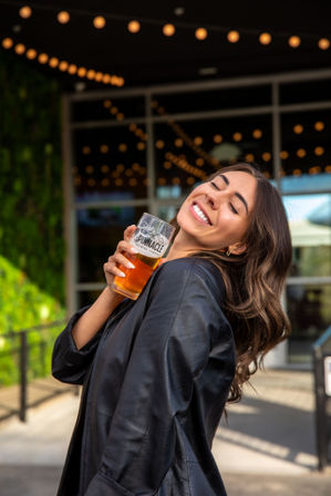 Smiling woman in a black leather jacket holding a pint of craft beer on a string-lit outdoor patio