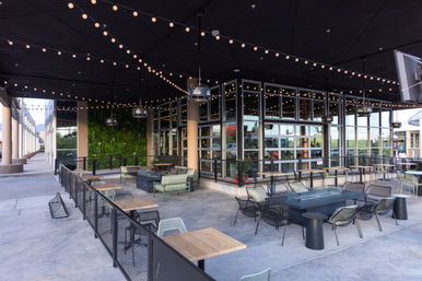 Covered outdoor patio at a modern shopping center — al fresco restaurant seating with twinkling string lights, lounge sofas, metal tables and chairs, a green living wall and glass storefront.