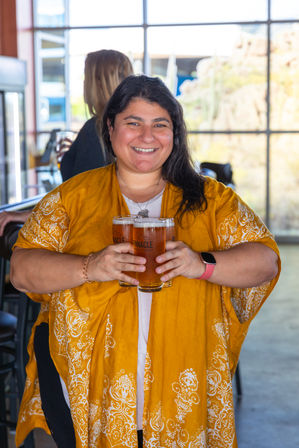 Smiling woman in a mustard yellow patterned shawl holding two pints of amber beer inside a sunlit brewery taproom with large windows.