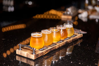 Flight of five craft beers in tasting glasses on a wooden paddle at a glossy bar counter, warm bokeh string lights in the background